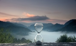 A hourglass (with falling sand) on a wooden wet table with sea of fog and mountain range silhouette in sunrise time