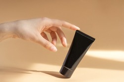 Woman's finger touches the mock-up of black tube of cream. Brown background with rays of sunlight. Packaging for cosmetics. Skin care concept.