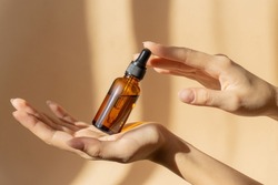 Mockup of glass bottle with dropper lid in female hands. Amber-colored container with cosmetic product, serum on brown background in rays of sunlight. Concept of beauty