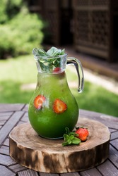 on a brown wooden table on a wooden log cabin is a jug of punch. it is a fresh green color. it consists of strawberries and spinach. you can see the strawberry halves in the jug. the drink is decorate
