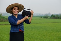 Asian female farmer holds radio receiver on shoulder. Listening to music at paddy field. Concept : Happy working along with music, news, information and advertisement from radio. Country lifestyle. 