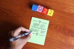 Top view image of left hand write text Happy Left Hander's Day on green paper on wooden table