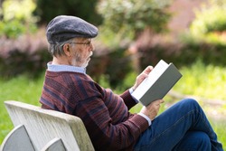Caucasian old man reads a book on the park bench. Concept of serene old age.