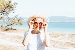 Happy senior woman smiling while holding the sunglasses. Retirement resting, new healthy habits, traveling and doing activities. Smiling and having fun outdoors