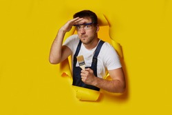 caucasian painter worker in safety glasses and uniform holding paint brush and looking forward through hole in yellow paper.