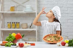 child making tasty delicious gesture by kissing fingers. little girl in chef hat and an apron cooking pizza in the kitchen.