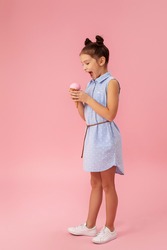 cute smiling little girl eating vanilla ice cream in a waffle cone on pink background. full length