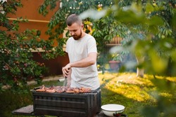 A young man in a white t-shirt with a beard makes a barbecue on the grill in the garden near his house. The guy fries pork kebabs on a metal grill.