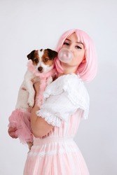 A girl with pink hair and in a pink dress inflates a chewing bubble and holds her dog in a pink outfit, isolated on a white background. A girl with pink hair and freckles blows gum.