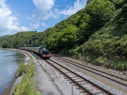 steam train on train track next to water and near a hill with trees and blue sky in Paignton, Devon, UK