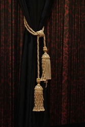 A solid black drape wrapped with a gold cord adorned with a gold tassel against the backdrop of a patterned black and red drape.