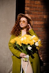 Close up portrait of happy stylish woman with red curly hair in a green coat with a bouquet of yellow tulips. Woman posing on camera and enjoying sunny spring time.