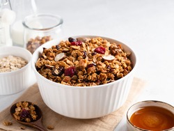 Granola (baked rolled oats with nuts and dried cranberries) in white ceramic bowl on beige table napkin, bottle of milk, honey bowl and oat flakes. Close up view. Healthy eating. Breakfast background