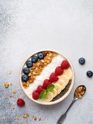 Muesli and yogurt bowl decorated with fresh blueberry, raspberry, banana slices, chia seeds, coconut shred. Healthy vegetarian breakfast or snack, tea spoon. Top view. Copy space. Grey background.