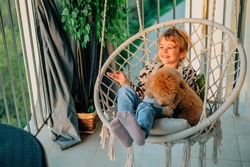happy little girl, child hugging with a smile her pet, poodle dog at home on the balcony in spring, summer in a cotton-fringed hammock chair at sunset. The animal is like a member of the family.