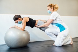 Caucasian female physical therapy professional at a clinic giving a pilates lift treatment with a therapy ball to a client with a face mask due to the covid 19 coronavirus pandemic.