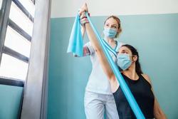 Professional Caucasian physical therapy woman at a clinic giving a Pilates stretch treatment with a rubber band to a client with a face mask due to the covid 19 coronavirus pandemic.