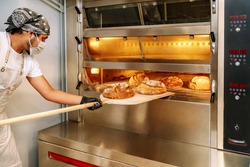 selective focus of Latino baker putting bread in the oven with gloves and a mask due to the 2020 covid19 coronavirus pandemic
