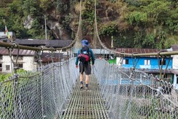 A young male trekker crossing a suspension bridge to a small village on the Annapurna circuit trail. Man wearing a blue hat, a backpack with a sleeping bag and sturdy boots.