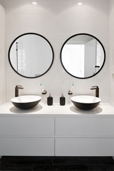 Symmetrical image of a bathroom with two sinks, two black faucets, two round mirrors with black frames and a white wooden cabinet

