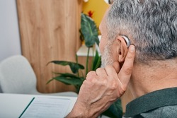 Hearing solutions for elderly deafness people. Older gray-haired man tunes his hearing aid behind the ear by pressing his finger on setting button while visit to hearing clinic