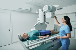 Male patient lying on bed while female nurse adjusting modern X-ray machine for scanning his leg or knee for injuries and fractures