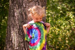 A little girl looking at her tie dye tee shirt- A little girl holding her new shirt up to her and looking at the tie dye design on it- A child is standing outside by a tree admiring her new shirt 