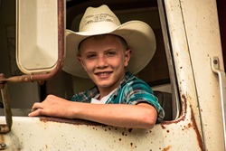 A young country boy with a cowboy hat on looking out the window of a old rusty vintage work truck- A happy, smiling teenager in the front seat of an old farm truck- A young man driving a old truck