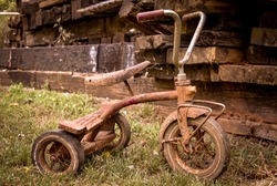 A old rusty vintage tricycle from the 1960s sitting outside with a pile of wood and timbers behind it- A child's metal toy bike  sitting outside 