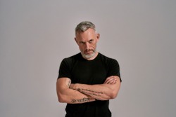 Strength. Middle aged man with crossed arms in black t shirt looking at camera while posing in studio over grey background. Front view. Horizontal shot