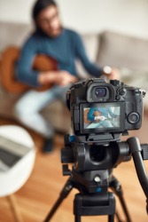 Guitar lessons. Young man sitting on sofa at home and teaching how to play guitar. Recording webinar at home. Learning music online. Focus on a camera. Stay home. E-learning. Music school online