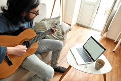 Young man sitting on sofa at home and learning how to play guitar, watching online course on laptop. Distance education. Main focus on laptop. Stay home. E-learning. Music school online
