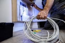 Close up shot of hand of aged electrician repairman in uniform working, fixing, installing ethernet cable in fuse box, holding flashlight and cable. Selective focus on hand and cable. Horizontal shot