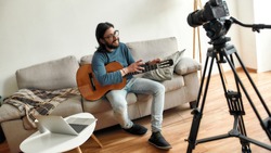 Music school online. Young man blogger sitting on sofa at home and teaching how to play guitar online. Recording video tutorial at home. Focus on a camera. Stay home. E-learning. Online education