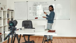 Javascript lesson. Young bearded male teacher wearing glasses pointing at whiteboard and teaching Javascript, giving lesson online. Recording video blog. Focus on a man. E-learning. Distance education