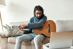 Young caucasian man sitting on sofa at home and adjusting acoustic guitar, watching guitar lesson online on laptop. Music school. Distance education. Focus on man. Stay home, self isolation. Elearning