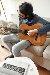 New skill. Young focused man sitting on sofa at home and playing acoustic guitar, watching online music course on laptop and practicing chords. Distance education. Focus on man. Stay home. E-learning