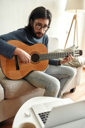 Learning a new instrument. Young focused man wearing glasses sitting on sofa at home and learning how to play guitar, watching online course on laptop. Distance education. Focus on a man. Stay home