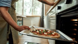 Cropped shot of man, professional cook making pizza at home. Man in apron putting raw pizza in modern oven for baking. Hobby, lifestyle. Selective focus. Side view. Web Banner