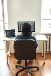 Back view of male web developer writing code while sitting at his workplace and working from home, writing code. Using desktop computer and laptop. Freelance and home office. Stay home, self isolation