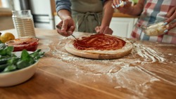 Cropped shot of couple making pizza together at home. Man in apron adding, applying tomato sauce on the dough while woman adding mozzarella cheese. Hobby, lifestyle. Selective focus. Web Banner