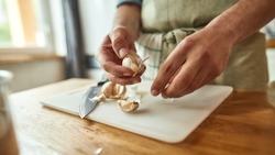 Close up of hands of man in apron peeling garlic while preparing a meal in the kitchen. Cooking at home concept. Selective focus on hands. Web Banner