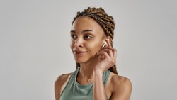 Listening music. Portrait of young attractive fitness woman wearing wireless earbuds and smiling, standing isolated over grey background, concept of sport, fitness and healthy lifestyle