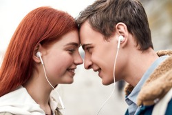Close up of happy couple of teenagers listening to music using the same pair of earphones, looking at each other with love. Technology, romance concept. Side view. Horizontal shot