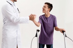 Cropped shot of male doctor shaking hands with cheerful disabled boy with cerebral palsy, taking steps using his walker isolated over white background. Children with disabilities and special needs
