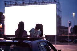 Rear view of two female friends sitting in the car while watching a movie in an open air cinema with a big white screen. Entertainment concept. Focus on people. Horizontal shot