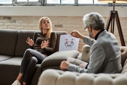 Depressed young caucasian woman talking about Rorschach test pictures with psychotherapist during appointment in office, selective focus. Psychotherapy concept