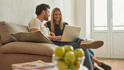Couple on the sofa discusses something funny and laughs, sitting near the balcony , white table with magazines and bowl of green apples unfocused on the front