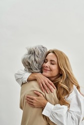 Loving grownup daughter hug embrace old mother feel grateful and thankful, isolated on white studio background. Caring adult child cuddle mature mom. Vertical narrow shot. Motherhood.