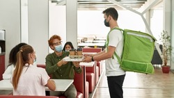 Male employee taking pizza and coffee from courrier during break with multiracial female colleagues at work. People wearing medical masks. Delivery at Covid-19 pandemic. Idea of rest and break at work